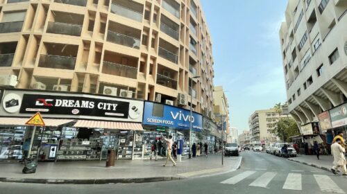 Strada commerciale di Dubai con negozi di elettronica, passanti e palazzi residenziali in luce diurna