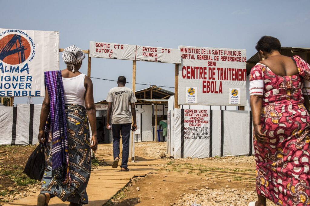 Tre persone camminano verso l'ingresso di un Centro di trattamento Ebola in Repubblica Democratica del Congo; cartelloni in francese e swahili indicano entrata, uscita e orari di visita
