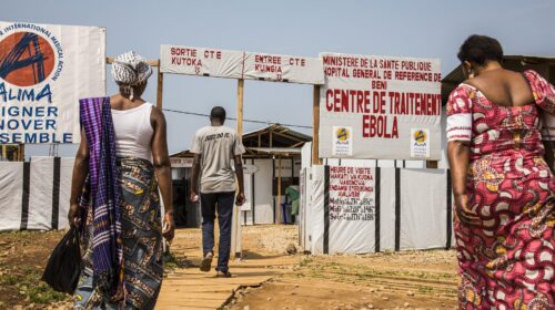 Tre persone camminano verso l'ingresso di un Centro di trattamento Ebola in Repubblica Democratica del Congo; cartelloni in francese e swahili indicano entrata, uscita e orari di visita
