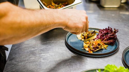 Mano di un cuoco che completa l'impiattamento di una tostada con verdure, fagioli neri e mais su un piatto blu in una cucina professionale